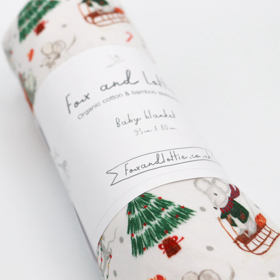 A rolled Christmas baby blanket by Fox and Lottie. Print has Christmas tree, mouse on sleigh, mouse wrapped in Christmas lights.