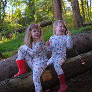 Two young children sitting on a fallen log in a wooded outdoor setting, wearing matching white Christmas-themed pyjamas with red and green Christmas prints and bright red welly boots.