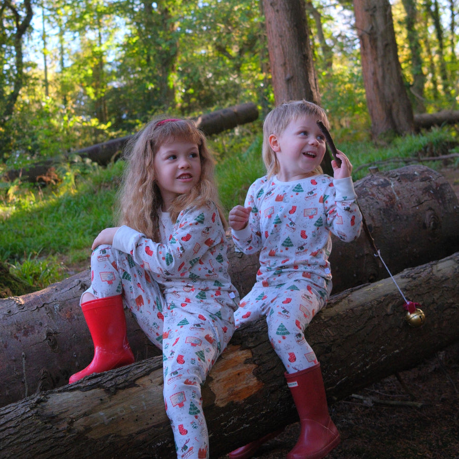 Two young children sitting on a fallen log in a wooded outdoor setting, wearing matching white Christmas-themed pyjamas with red and green Christmas prints and bright red welly boots.