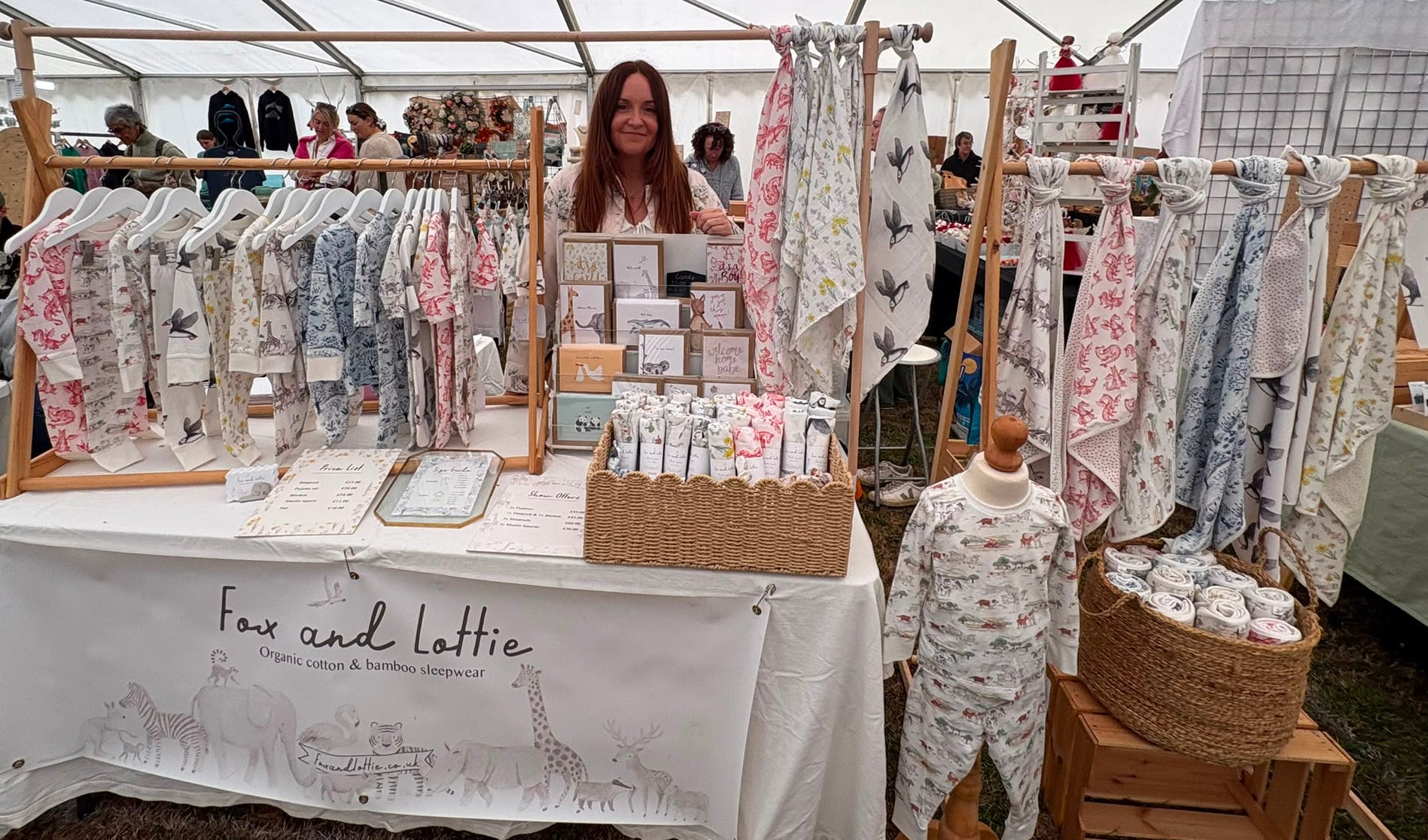 A market stall under a white tent displaying baby clothing and accessories from Fox and Lottie. The white tablecloth features animal illustrations and the brand name, with wooden racks holding baby garments and swaddles, and a mannequin wearing a printed onesie.